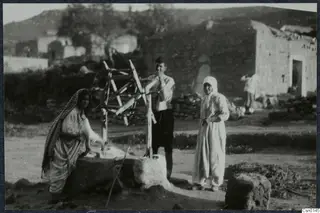 Three people beside a stone well with a wooden frame in a village street, with a stone building behind them