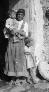 Woman in a headscarf holding a toddler in a doorway, with a small child standing beside her