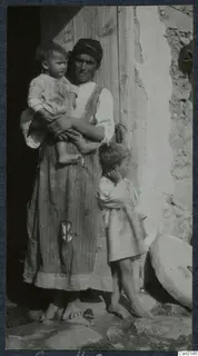 Woman in a headscarf holding a toddler in a doorway, with a small child standing beside her against a stone wall