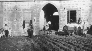 Stone building with an arched doorway and barred window, with people, donkeys and a dog in front of a ploughed field