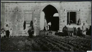 Men, children and a saddled donkey before a stone building with a pointed-arch doorway, beside a ploughed field