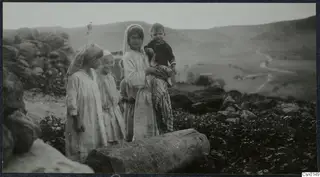 Children standing by a large cut stone block in a rocky field, with hills and a winding road behind