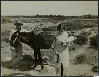 A man leading a dark donkey with a wicker basket on its back, and a woman carrying a bundle, on a dirt track
