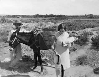 A man in a straw hat leads a donkey with a large wicker basket on its back, while a woman carries a white bundle on a dry track.