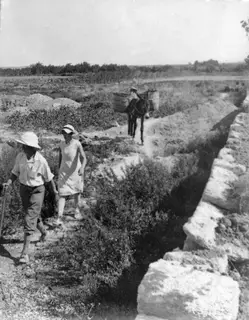 Man and woman walking along a dirt path beside a stone wall, with a pack donkey carrying baskets behind them
