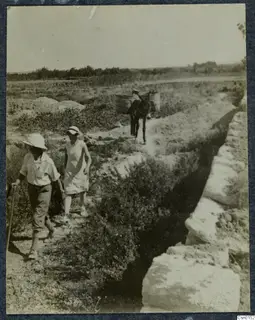 A man and a woman walk beside a stone wall, with a pack donkey carrying baskets on the path behind them