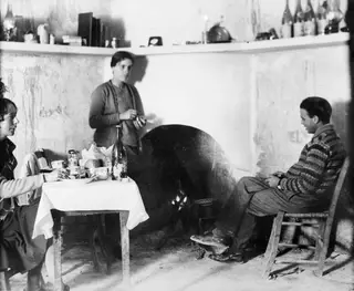 Three people in a whitewashed room beside a hearth, with a table laid for a meal