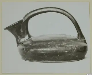 Dark-surfaced pottery jug with a high strap handle and a spout, photographed from the side on a plain background