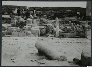 Excavated stone walls at Salamis, Famagusta, with a fallen column drum in the foreground and people standing among the ruins.