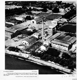 Aerial view of a winery with a tall chimney and sheds beside the sea