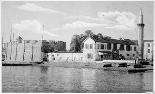 Waterfront view with a stone fortification, a white two-storey building, a minaret and small boats on the shore