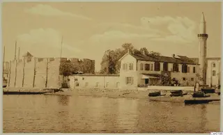 Waterfront with a stone fortification, a two-storey building and a minaret, with small boats drawn up on the shore