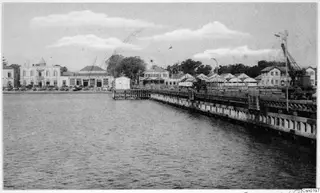 Quay with a crane and white buildings along a waterfront, seen across open water