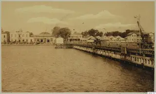 Waterfront quay with a crane and two-storey buildings beside calm water, shown in a brown-toned photograph