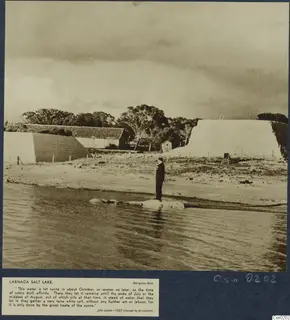 Man standing in shallow water at the edge of Larnaca salt lake, with long white-faced structures on the shore