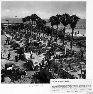 Seafront promenade with palm trees, cafe tables, parked cars, bicycles and crowds, with boats moored along the water.