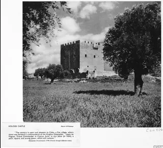 Kolossi Castle, a square stone tower with battlements, seen across a grassy field with trees on either side.