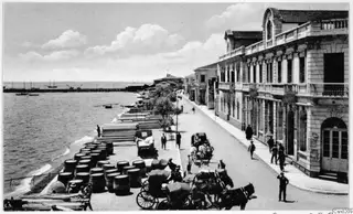 Waterfront street with stacked barrels and timber along the quay, horse-drawn carriages, and stone buildings with balconies
