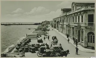 Seafront street with horse-drawn carts, stacked barrels along the quay, a long pier and stone buildings with balconies