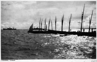 Long jetty at Limassol lined with bare masts of moored boats, a sailing boat and steamship offshore, and figures standing on the pier.