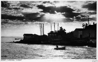 Sunset over the shore at Limassol, with masted boats and buildings along the water and one person rowing a small boat.