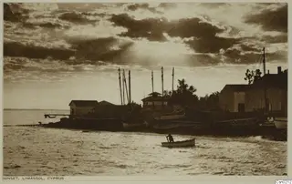 Sunset over the shore at Limassol, with boats with masts drawn up by white buildings and one person rowing a small boat.