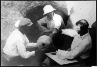 Three men seated by a rock opening, examining a rounded pale object and small items on a board
