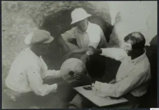 Three men handling a rounded object beside a dark rock-cut opening, one seated at a small board