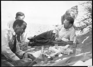 Four men relaxing on sand beside bottles, glasses and scattered food, one reclining in round spectacles.