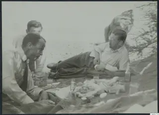Three men resting on sand beside bottles, glasses and food spread on a cloth.