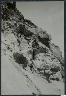 A man in a light shirt and shorts stands beside a rocky outcrop on a steep, pale slope.