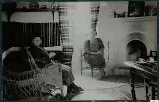 Two women seated in a room beside an arched fireplace, with a small table holding cups and saucers.