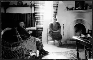 Two women seated in a room with a fireplace, a wicker armchair and a small table with cups and saucers