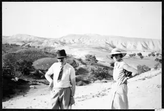Two people standing on a pale dirt slope, with eroded hills and scattered trees behind them.