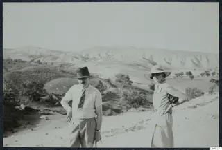 A man and a woman stand on a dusty track in front of pale hills, scrubby bushes and small trees.