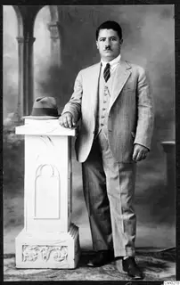 Studio portrait of a man in a pinstriped suit leaning on a white pedestal beside a hat