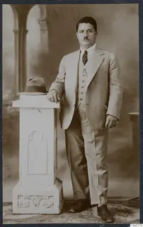 Studio portrait of a man in a pinstriped suit resting his arm on a white pedestal with a hat on top