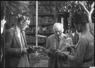 Three men handling small objects outdoors in front of stacked shelves, with a tree at the right edge and two more men behind.