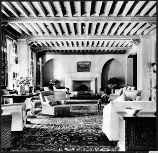 Living room interior with a beamed ceiling, arched alcoves, a fireplace and upholstered armchairs and sofas on a patterned carpet.