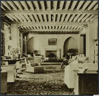 Furnished room in Nicosia with a beamed timber ceiling, arched alcoves, a fireplace, armchairs, a table and a framed picture.
