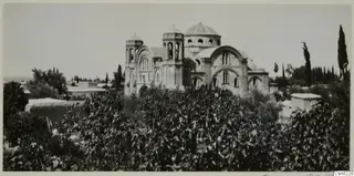 Domed church with bell towers and arched windows, seen above dense shrubs, with cypress trees at the right