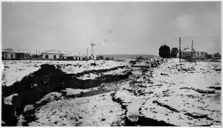 Deep eroded gully across flat ground, with low white houses, a windmill and trees behind and hills in the distance at Nicosia.