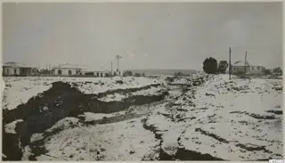 Deep eroded gully in the foreground, with white buildings, a windmill, trees and low hills beyond at Nicosia.