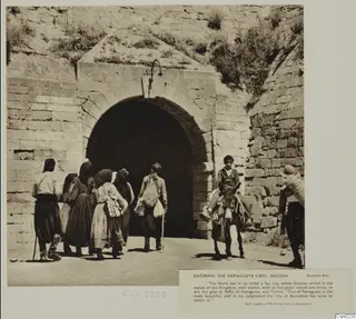 People walk toward a stone-arched city gate, with a rider on a donkey at the right edge