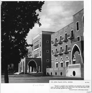 The Ledra Palace Hotel in Nicosia, with arched entrances and balconies, seen beside a large tree on the left.