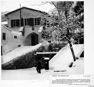 Paphos Gate, Nicosia, under snow, with a figure in a dark coat walking past a stone wall and a post box.