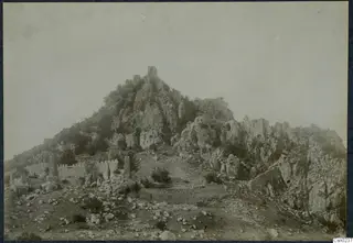 Rocky mountain peak with ruined crenellated walls and a tower, stones scattered in the foreground