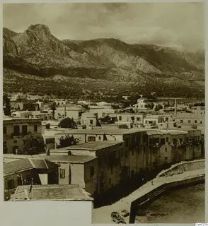 Flat-roofed houses of a town below steep mountains, with a harbour quay and small boats at the lower right