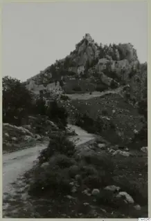 Dirt road through low bushes below a rocky mountain peak with ruined stone walls in the Kyrenia Mountains