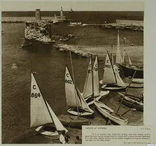 Sailing dinghies with numbered sails drawn up on a harbour quay at Kyrenia, with a ruined stone tower and mole behind.